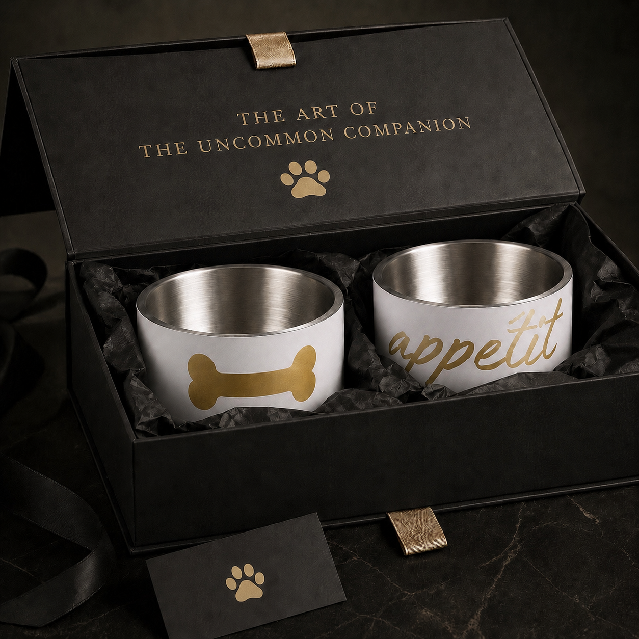 A black gift box lid open, displaying two ivory dog bowls with stainless steel interiors. Bone image and appetite dog bowl set. 