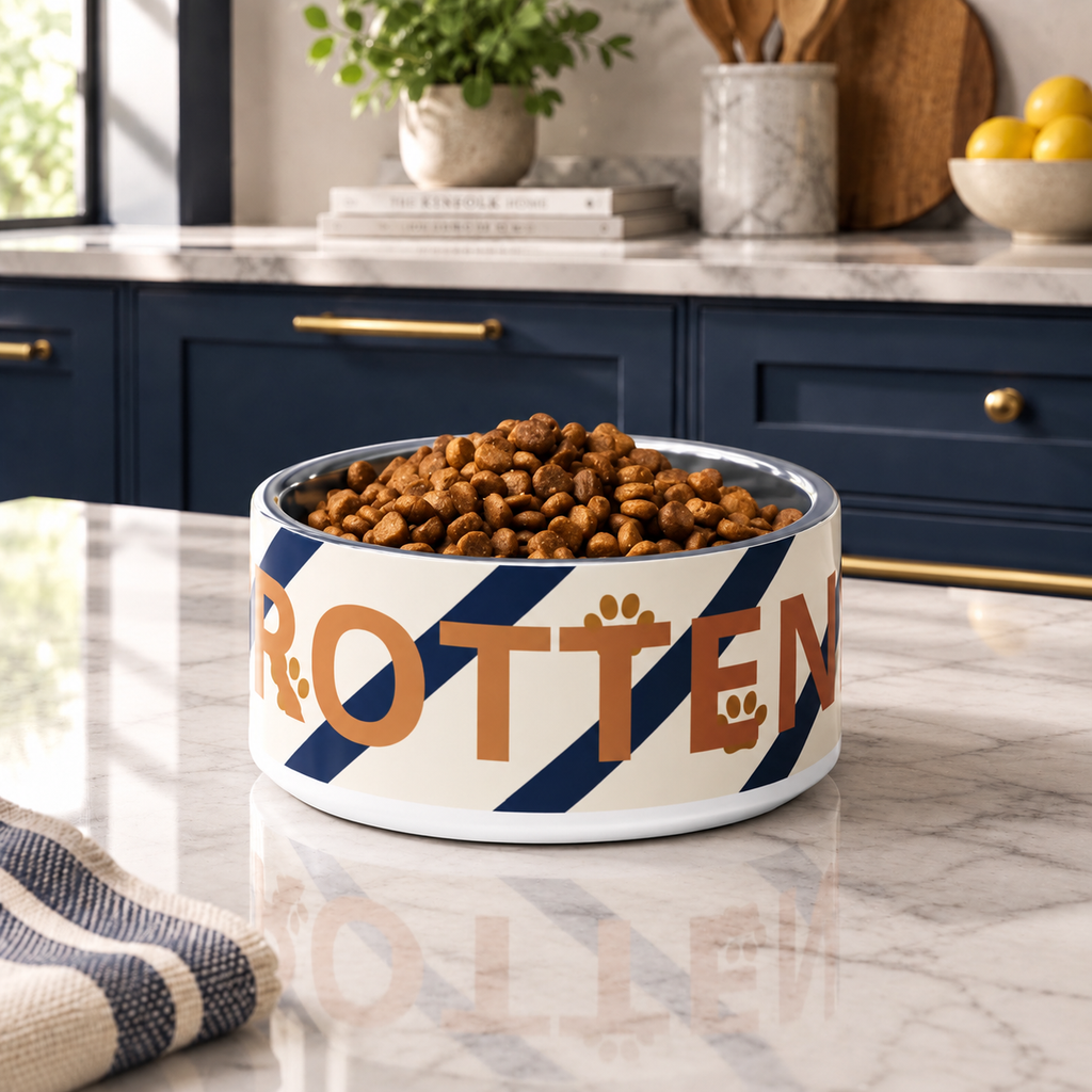 A ivory stainless steel dog bowl with navy blue diagonal stripes & the word 'ROTTEN' in gold, set on a marble kitchen countertop.