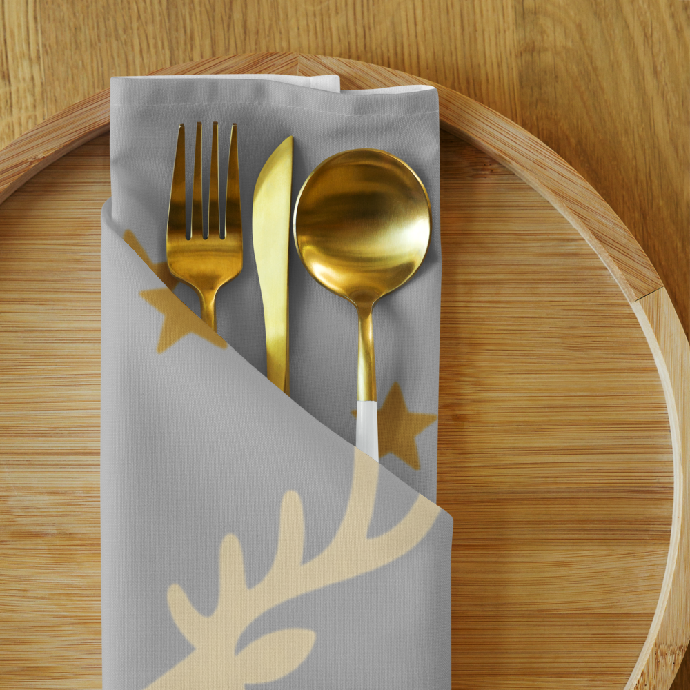 A folded silver cloth napkin with gold reindeer antler and star patterns, holding gold-toned flatware on a round wooden tray.