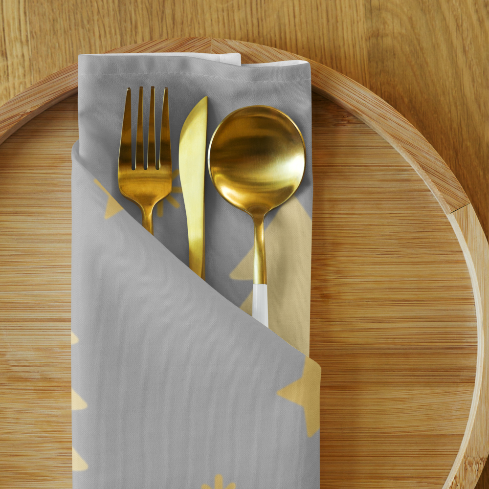 A folded sliver napkin with gold stars and trees, patterns hold a gold fork, gold knife, and gold spoon on a round wooden tray.