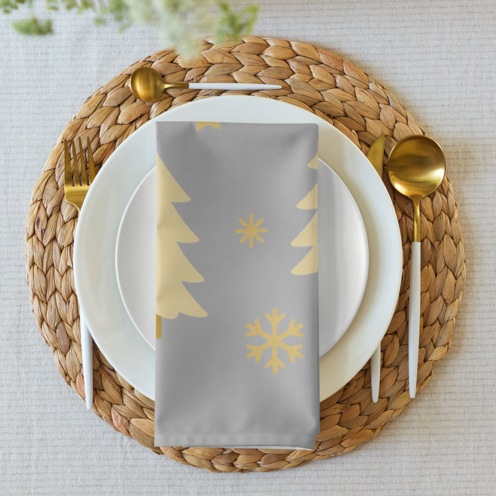A styled table setting featuring a silver cloth napkin with gold printed Christmas trees and snowflake motifs, layered over white plates with gold-toned cutlery.