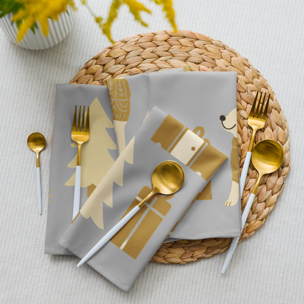 Silver cloth napkins with gold holiday-themed illustrations, styled on a woven placemat with gold flatware and a white tablecloth.