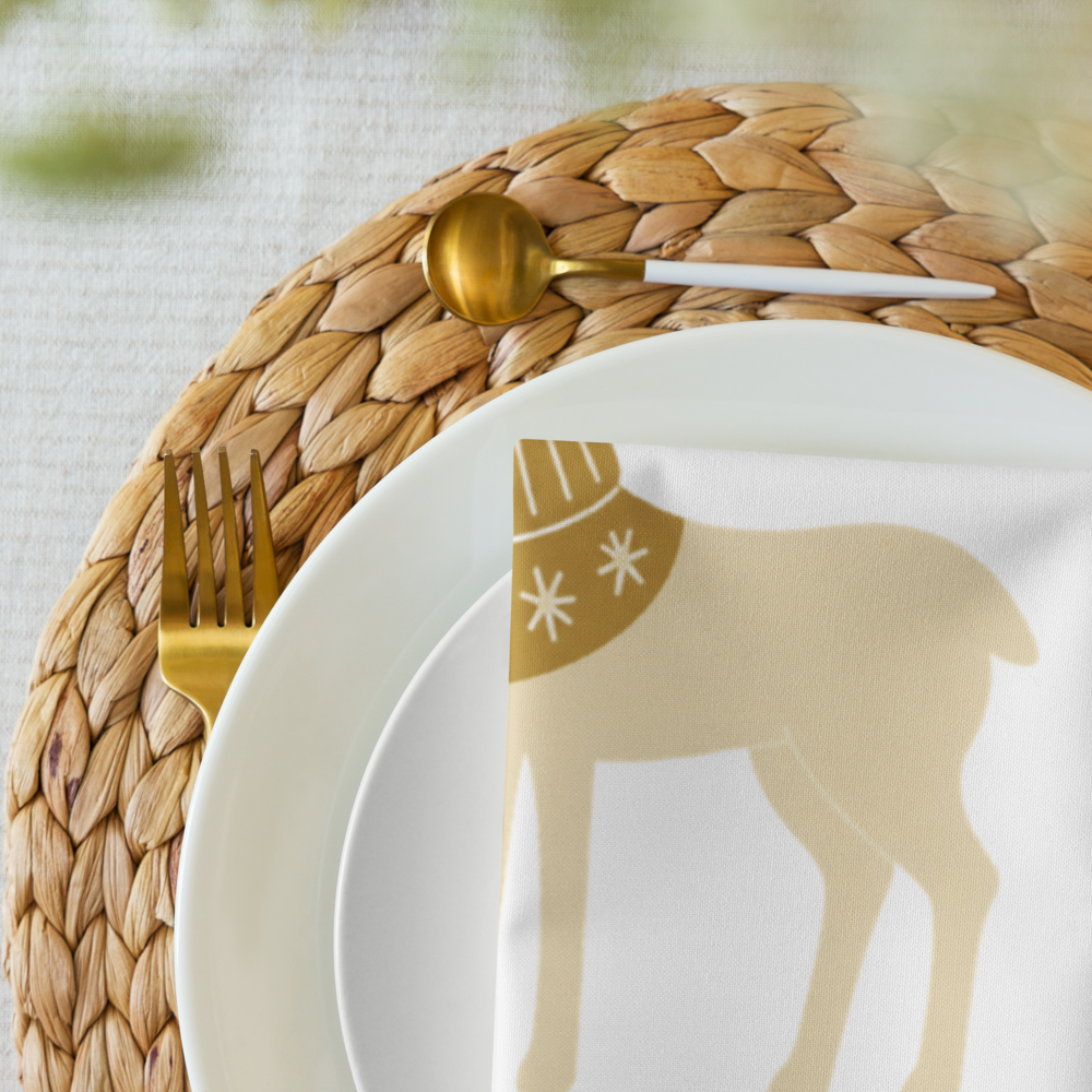 A close-up of a white dinner plate with a gold reindeer silhouette and white snowflake details, styled on a woven tan placemat with gold-toned cutlery and a gold spoon.