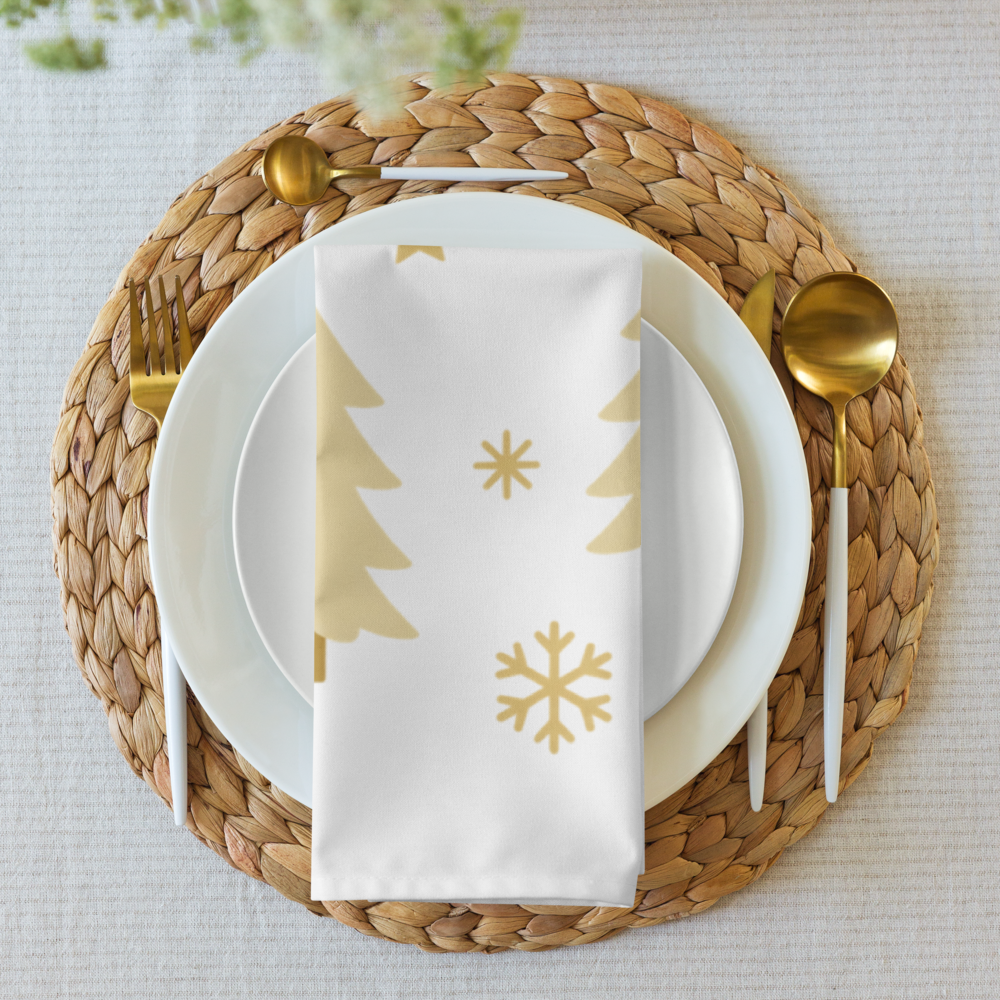 A styled table setting featuring a white cloth napkin with gold printed Christmas tree and snowflake motifs, layered over white plates with gold-toned cutlery.