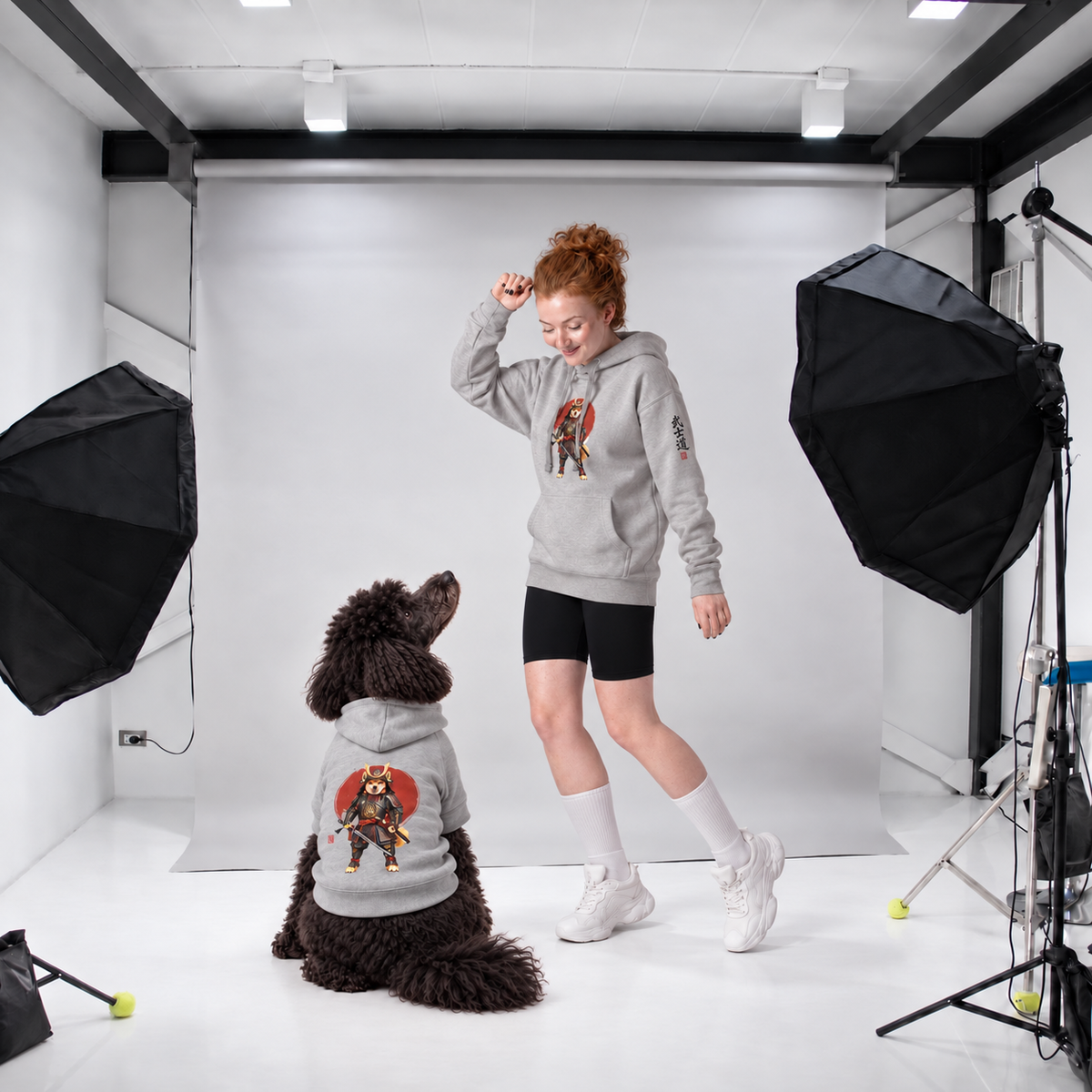 A woman and a black poodle modeling matching light gray hoodies with a Samurai warrior dog graphic print, set against a white studio backdrop.