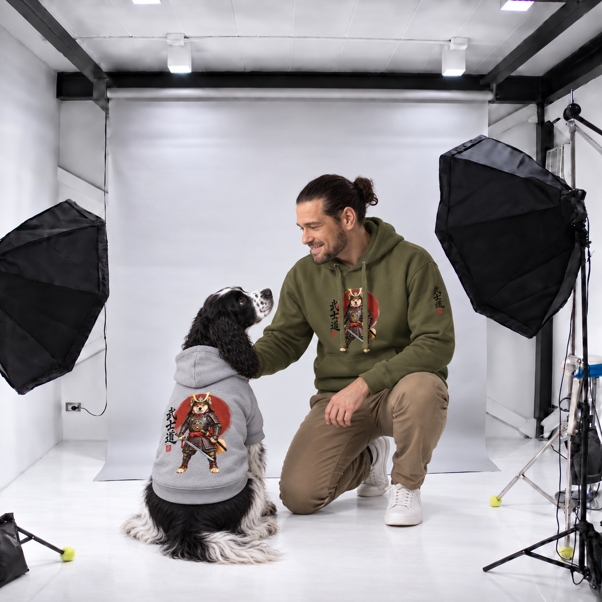 A man kneels on the floor next dog wearing matching graphic hoodies with a samurai dog design. The man hoodie military green the dog hoodie is gray.