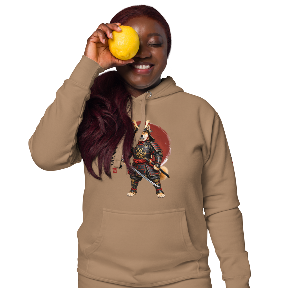 A smiling woman wearing hoodie featuring a graphic of a dog dressed as a samurai warrior. She is holding a yellow citrus fruit up to her eye.
