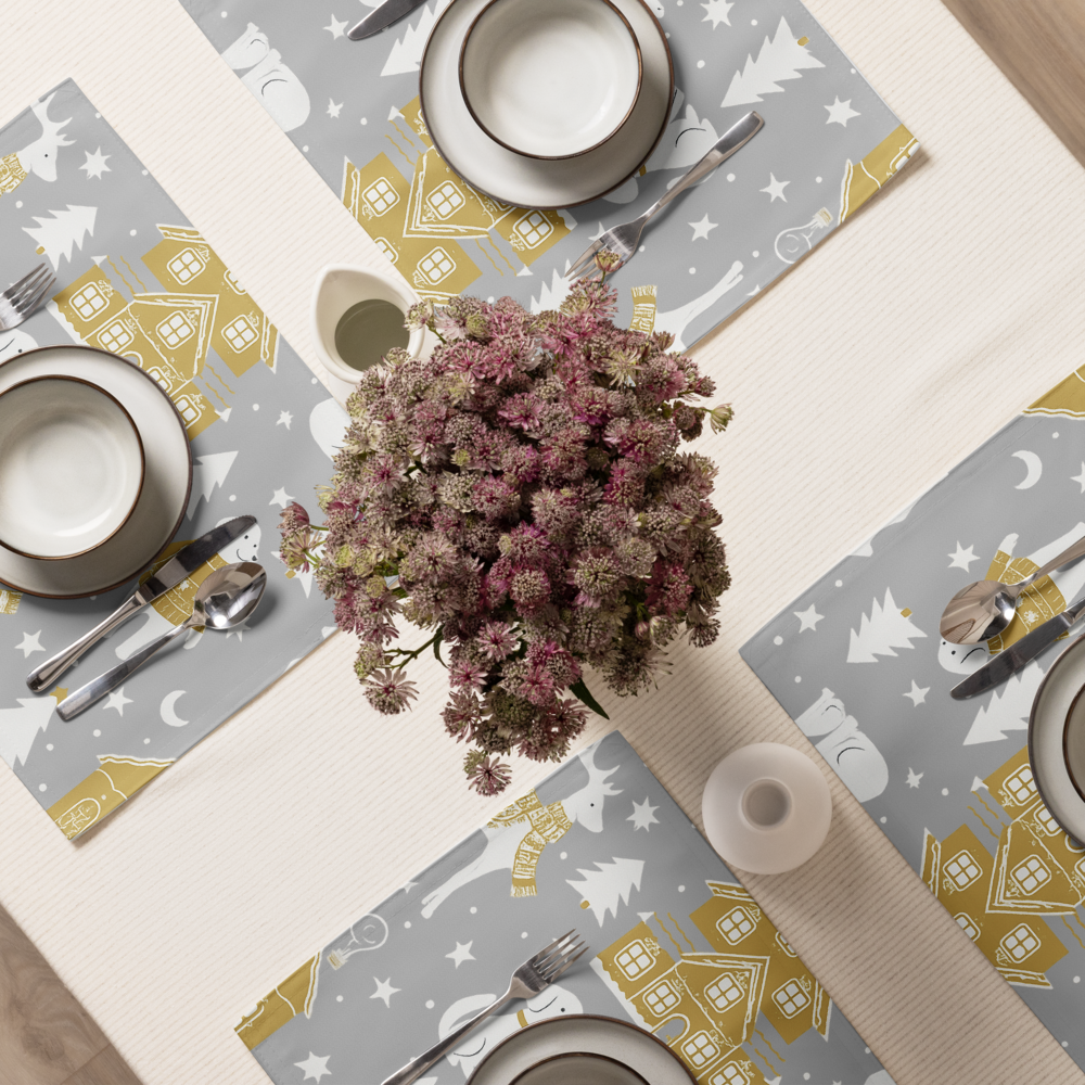 A styled dining table top viewed from above, featuring silver placemats with a winter holiday print of dogs, pine trees, stars, and gold houses, set with white plates.