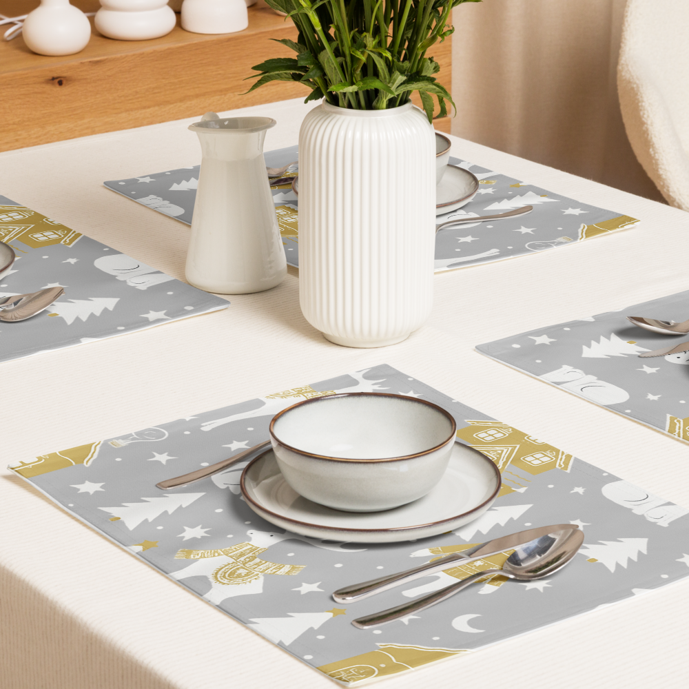 A styled dining table setting featuring silver placemats with a gold and white winter holiday pattern, including dog, pine trees, stars, and Christmas trees. The placemats are arranged on a white tablecloth alongside a white ribbed ceramic vase with green leaves, a small white pitcher, and white ceramic bowls and silver cutlery.